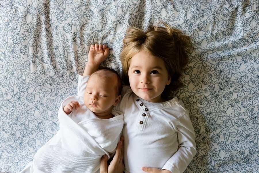 Un bebé envuelto en una manta blanca yace junto a un niño mayor de largo pelo castaño claro sobre una sábana estampada, ambos en paz. La escena es tranquila y atemporal, como capturada por reglas tácitas, como un procedimiento sumario de amor familiar.