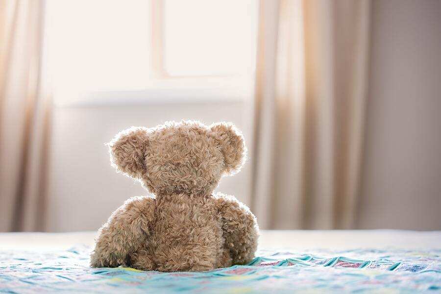 Un osito de peluche marrón está sentado en una cama frente a una luminosa ventana con las cortinas echadas.