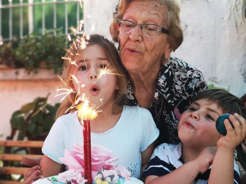 Durante un momento especial al aire libre, una anciana y dos niños pequeños comparten una tarta con una vela brillante. La escena capta la calidez familiar, perfecta para resaltar la importancia del régimen de visitas abuelos. Durante un momento especial al aire libre, una anciana y dos niños pequeños comparten una tarta con una vela brillante. La escena capta la calidez familiar, perfecta para resaltar la importancia del régimen de visitas abuelos.