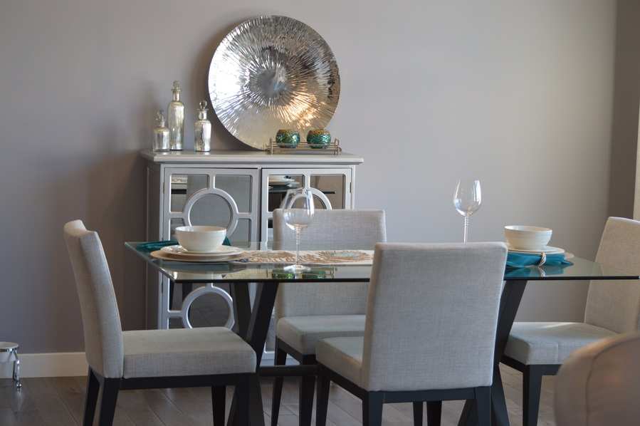 Un comedor moderno en una vivienda familiar presenta una mesa de cristal, cuatro sillas tapizadas en beige, cubiertos con cuencos y vasos, y un aparador con espejos rematado con objetos decorativos contra una pared gris claro.