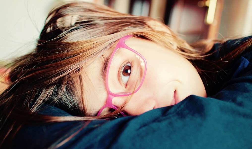 Una persona de pelo castaño largo y gafas rosas está tumbada con la cabeza apoyada en el brazo, mirando a la cámara. El fondo difuminado le da un aire tranquilo, como si se detuviera a reflexionar sobre unos gastos extraordinarios.