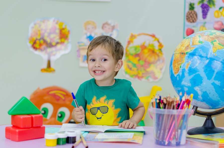 Un niño está sentado en una mesa, sonriente, sosteniendo un bolígrafo sobre un libro abierto. Sobre la mesa hay lápices de colores, pintura, billetes de gastos extraordinarios y bloques de construcción. Detrás de él hay dibujos y un globo terráqueo.