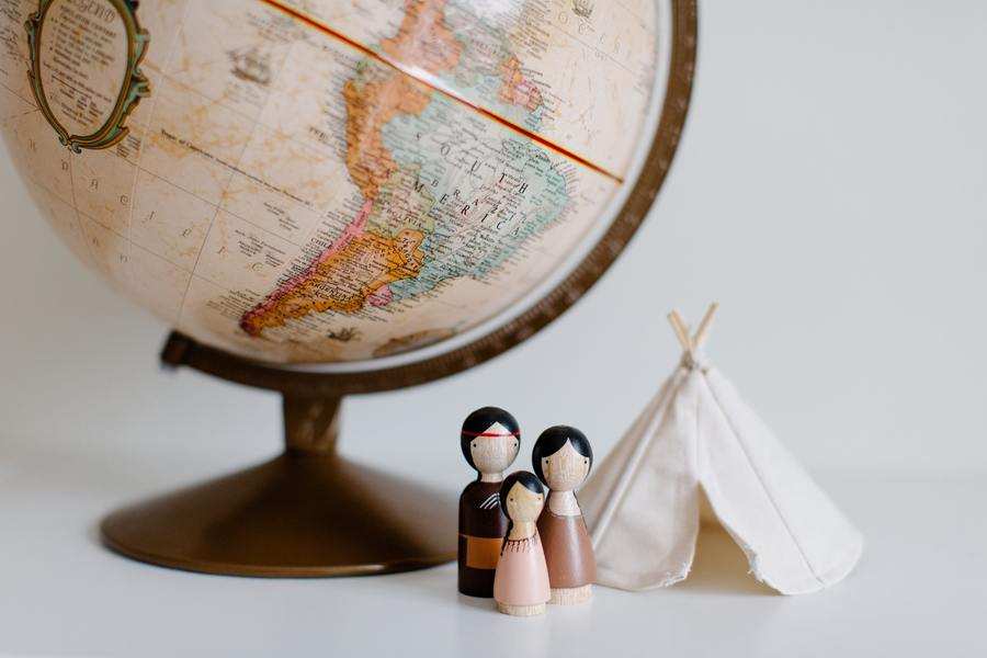 Un globo terráqueo de sobremesa junto a tres muñecos de madera y un pequeño tipi de tela blanca evocan temas familiares y de convivencia sobre un fondo sencillo y liso.