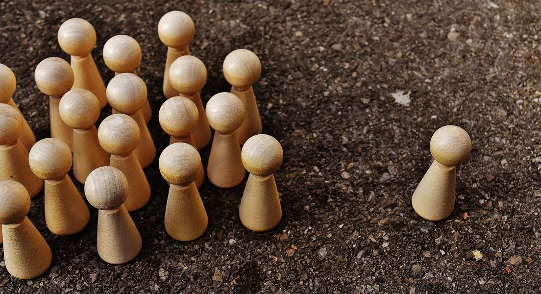 Varios peones de madera se agrupan sobre una superficie áspera y oscura, mientras que un solo peón -que simboliza el acoso laboral- se encuentra solo, a cierta distancia del grupo. Varios peones de madera se agrupan sobre una superficie áspera y oscura, mientras que un solo peón -que simboliza el acoso laboral- se encuentra solo, a cierta distancia del grupo.