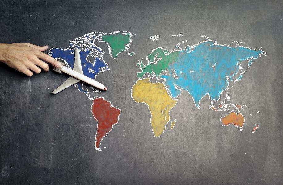 Una mano sostiene una maqueta de avión sobre un mapamundi de pizarra, con los continentes coloreados de forma diferente. El avión planea sobre Norteamérica, mientras el mapa de tiza blanca crea un exequátur de fronteras y destinos. Una mano sostiene una maqueta de avión sobre un mapamundi de pizarra, con los continentes coloreados de forma diferente. El avión planea sobre Norteamérica, mientras el mapa de tiza blanca crea un exequátur de fronteras y destinos.