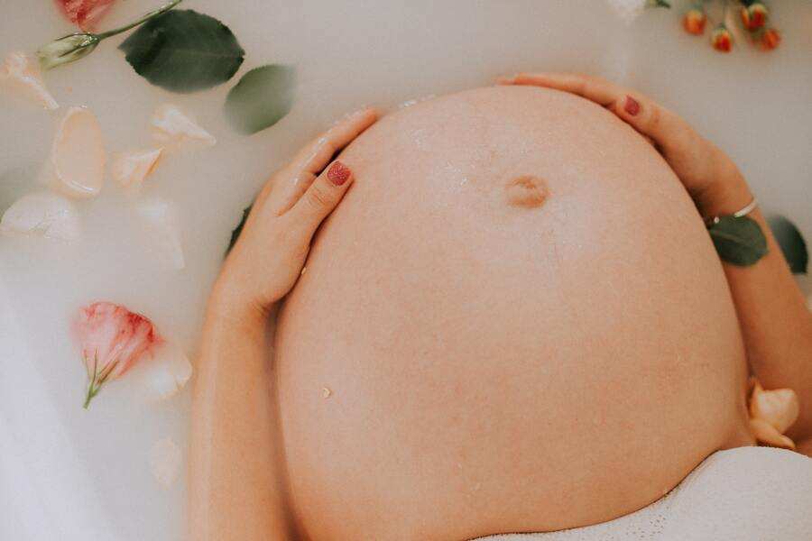 Primer plano del vientre de una mujer embarazada durante la gestación subrogada, tumbada en un baño de leche, con las manos apoyadas en el vientre y pétalos de flores flotando en el agua.