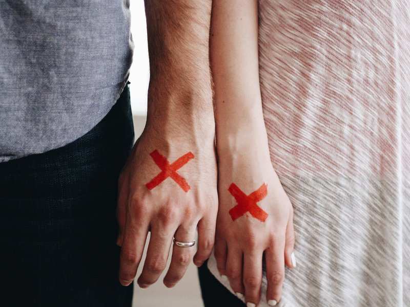 ¿es necesario acudir a la vía judicial en caso de divorcio? Dos personas están una al lado de la otra con grandes X rojas pintadas en el dorso de la mano, símbolo de divorciarse sin pasar por el juzgado. Uno lleva una camisa gris y un anillo; el otro, un top de rayas claras. Ambas tienen las manos relajadas y hacia abajo.