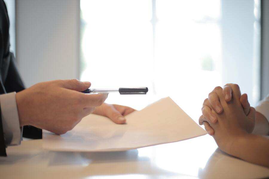 Una persona de traje sostiene un bolígrafo y un documento hacia otra con las manos juntas sobre la mesa, sugiriendo una firma o discusión de papeleo, posiblemente sobre cómo divorciarse sin pasar por el juzgado.