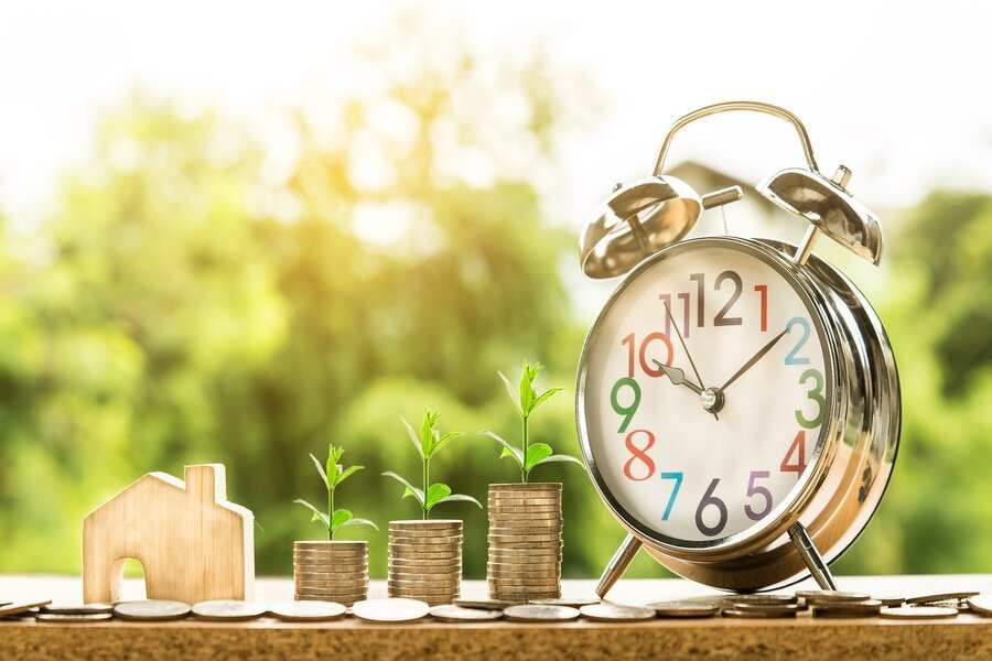 Un reloj despertador de plata, pilas de monedas de las que crecen pequeñas plantas que simbolizan la compensación económica por razón de trabajo, monedas sueltas y una pequeña maqueta de casa de madera se exhiben sobre una superficie con fondo verde difuminado. Un reloj despertador de plata, pilas de monedas de las que crecen pequeñas plantas que simbolizan la compensación económica por razón de trabajo, monedas sueltas y una pequeña maqueta de casa de madera se exhiben sobre una superficie con fondo verde difuminado.