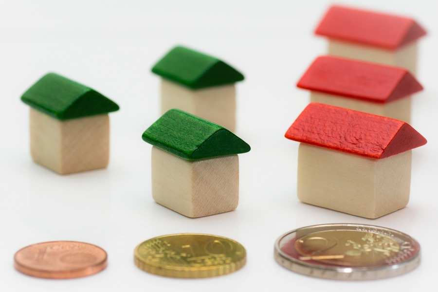 Cuatro pequeñas maquetas de casas de madera, dos con tejados verdes y tres con tejados rojos, están dispuestas en el fondo. En primer plano, tres monedas -de euro y de céntimo- sugieren una compensación económica por razón de trabajo sobre una superficie blanca.