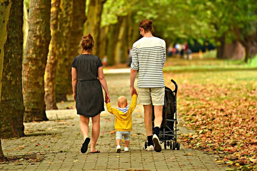 Dos adultos caminan cogidos de la mano con un niño por el sendero de un parque arbolado. Un cochecito está cerca mientras caminan entre las hojas caídas, quizás reflexionando sobre si extinguir la pareja de hecho es el paso correcto para su familia.