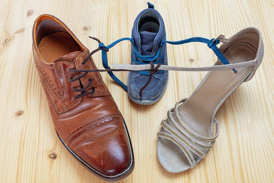 Un zapato de vestir marrón de hombre, una zapatilla de deporte azul de niño y una sandalia de tacón beige de mujer descansan uno junto al otro sobre una superficie de madera, la zapatilla azul atada a las otras, un sencillo símbolo de conciliación familiar.