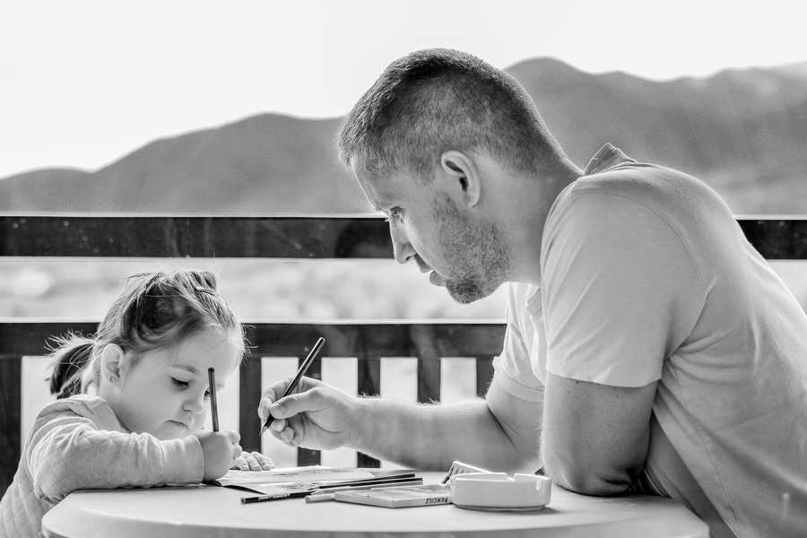 Un hombre y una niña, disfrutando de la custodia compartida progresiva, se sientan en una mesa redonda al aire libre con las montañas de fondo. Ambos sostienen bolígrafos y se concentran en dibujar o escribir juntos, rodeados de material artístico.