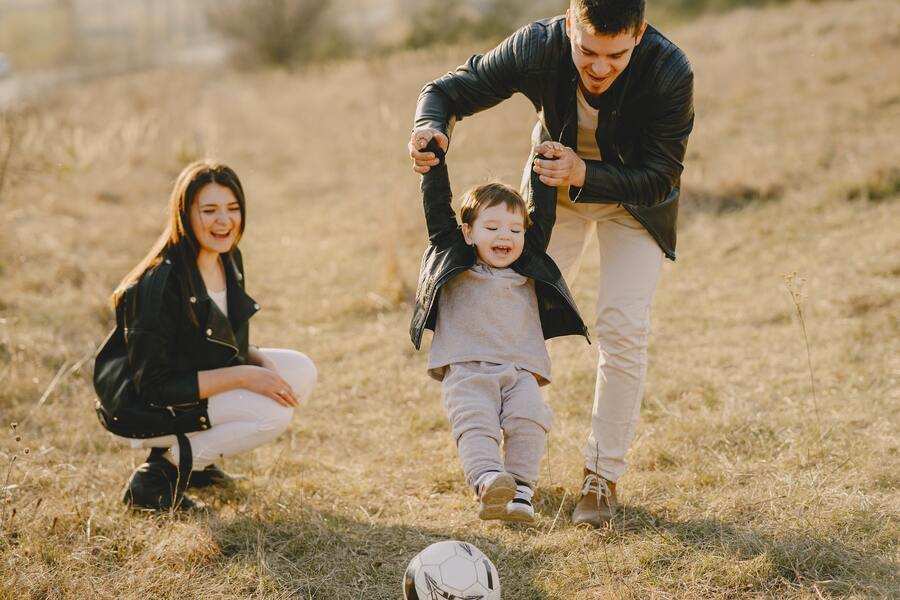 Un hombre sujeta a un niño pequeño por los brazos, levantándolo del suelo, mientras una mujer se arrodilla cerca. Están al aire libre en el césped con un balón de fútbol delante, compartiendo risas y tiempo de calidad como parte de su plan de parentalidad. Un hombre sujeta a un niño pequeño por los brazos, levantándolo del suelo, mientras una mujer se arrodilla cerca. Están al aire libre en el césped con un balón de fútbol delante, compartiendo risas y tiempo de calidad como parte de su plan de parentalidad.