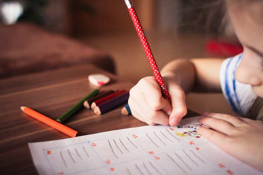 Un niño con un lápiz de lunares rojos escribe en una hoja de ejercicios en una mesa de madera, trabajando en silencio mientras los lápices de colores y una goma de borrar descansan cerca, capturando un momento reflexivo perfecto para ilustrar un plan de parentalidad en acción.