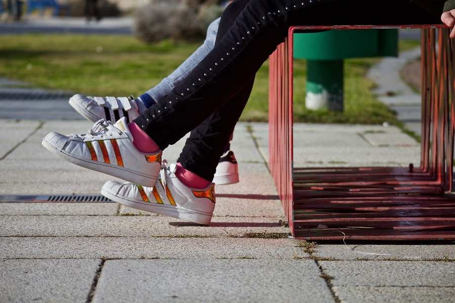 Dos personas se sientan en un banco de metal rojo al aire libre, con la parte inferior de las piernas y los pies a la vista, tal vez discutiendo temas como a qué edad pueden decidir los hijos con quién quieren vivir tras el divorcio. Ambos llevan zapatillas de deporte y pantalones negros; la hierba asoma por detrás del suelo pavimentado.