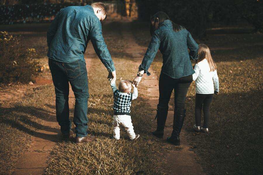 Dos adultos y dos niños pequeños caminan por un sendero de un parque. Los adultos llevan de la mano a un niño pequeño, mientras un niño mayor camina un poco por delante.