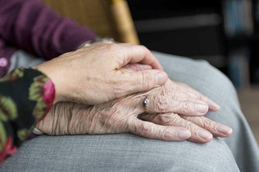 La mano de una persona joven descansa suavemente sobre la de un pariente mayor, con las manos cruzadas sobre el regazo. El calor entre ellos evoca la comodidad de compartir alimentos entre parientes sentados juntos en el interior.