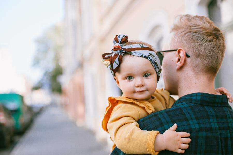 Un bebé con una cinta en la cabeza y un conjunto amarillo es sostenido por un adulto de pelo rubio corto y gafas, de pie en la acera de una ciudad, tal vez explorando las realidades de la custodia compartida y de vivir en el extranjero.