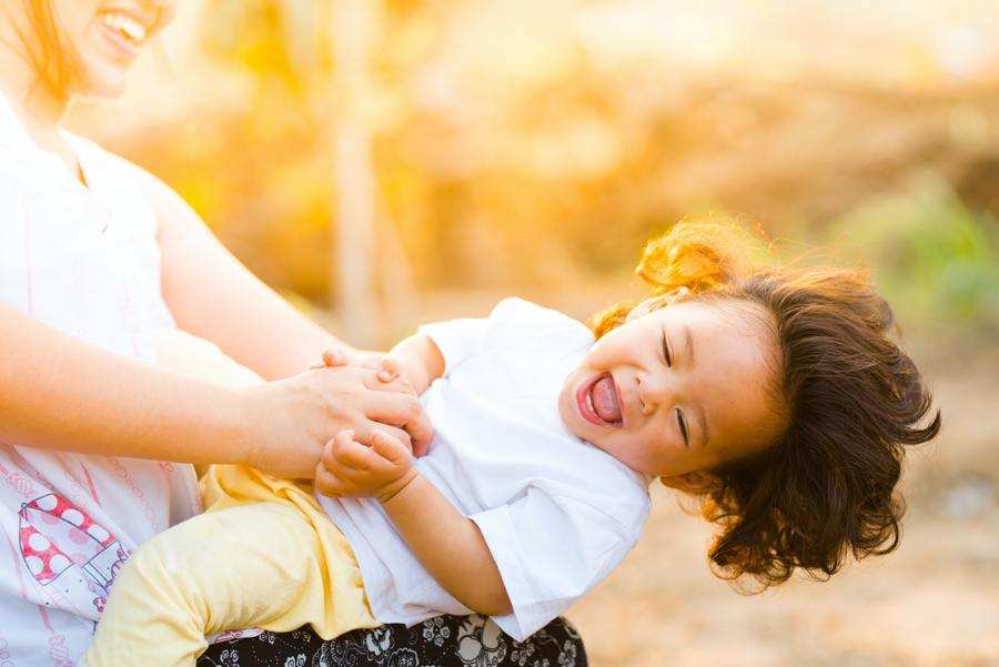 Un adulto, posiblemente un Coordinador Parental, sostiene en brazos a un niño sonriente con el pelo largo al aire libre en un entorno soleado. El niño, vestido con camisa blanca y pantalones amarillos, ríe alegremente mientras se reclina hacia atrás.