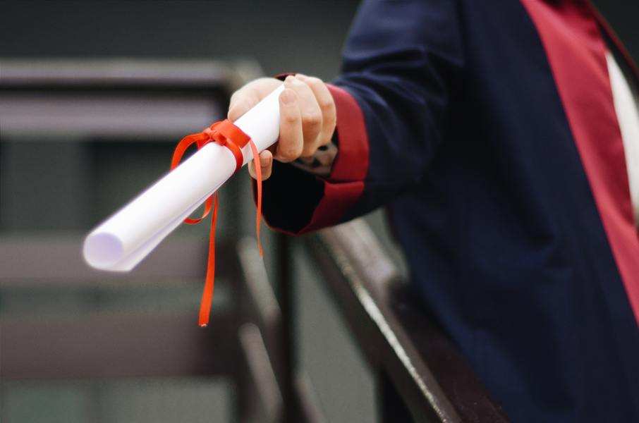 Una persona con toga académica oscura sostiene un diploma enrollado y atado con un lazo rojo.