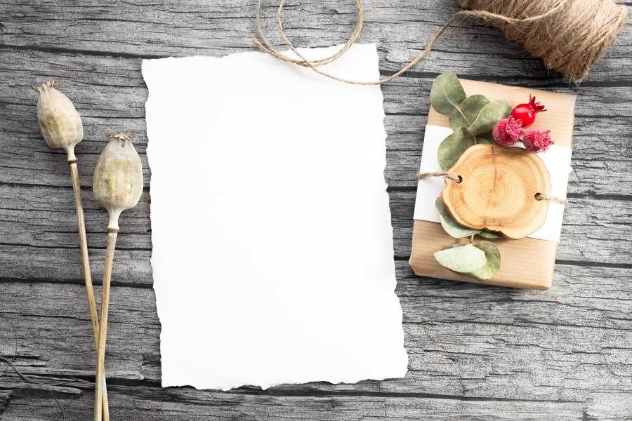 Una hoja en blanco de papel blanco rasgado sobre una superficie de madera, junto a vainas secas de adormidera, una tarjeta de madera decorada con hojas y flores rojas y una bobina de hilo: una disposición que evoca la formalidad del papeleo de la sucesión intestada.