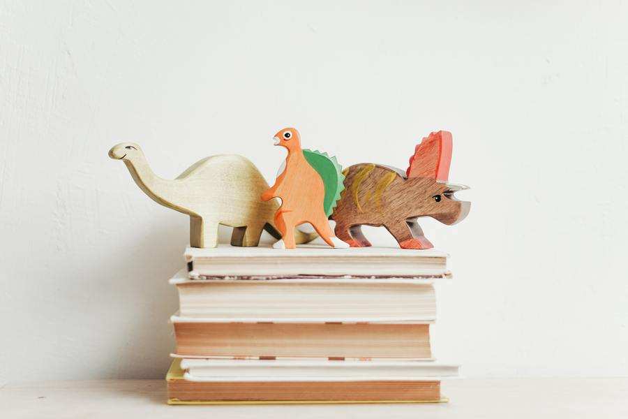 Tres figuras de dinosaurios de madera en beige, naranja y marrón se sitúan sobre una pequeña pila de libros de tapa dura sobre un fondo blanco liso, perfecto para decorar un espacio de entregar ropa a hijos tras un divorcio.