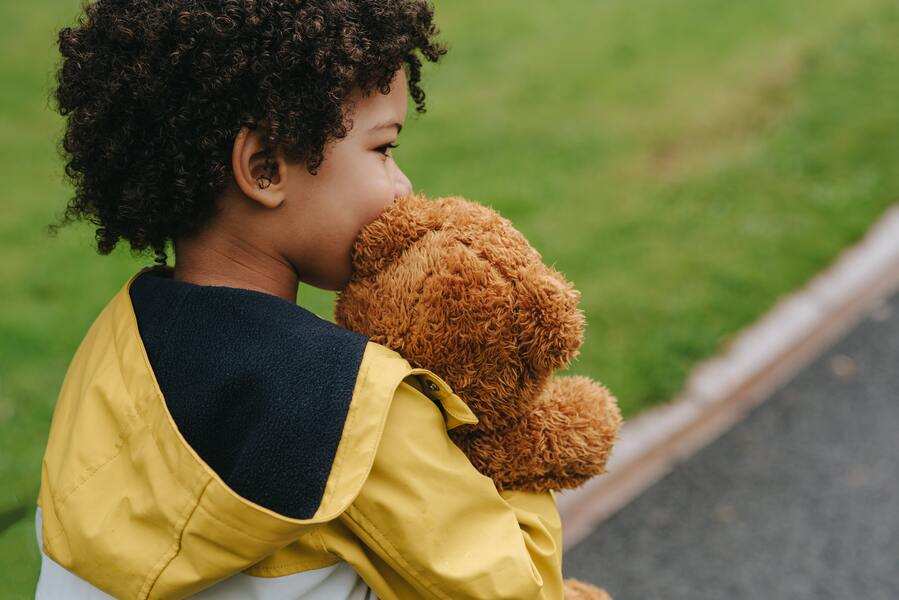 Un niño con un chubasquero amarillo sostiene un osito de peluche marrón cerca de la cara mientras está al aire libre en una zona de hierba junto a un camino, como si reflexionara sobre cómo la vida puede cambiar o modificar la pensión de alimentos.