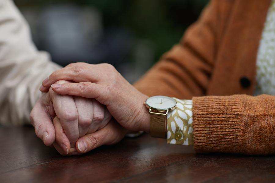 Una persona con jersey marrón y reloj de pulsera sujeta suavemente la mano de otra a través de una mesa de madera, simbolizando el apoyo cuando se habla de temas como el poder preventivo. El fondo está borroso. Una persona con jersey marrón y reloj de pulsera sujeta suavemente la mano de otra a través de una mesa de madera, simbolizando el apoyo cuando se habla de temas como el poder preventivo. El fondo está borroso.