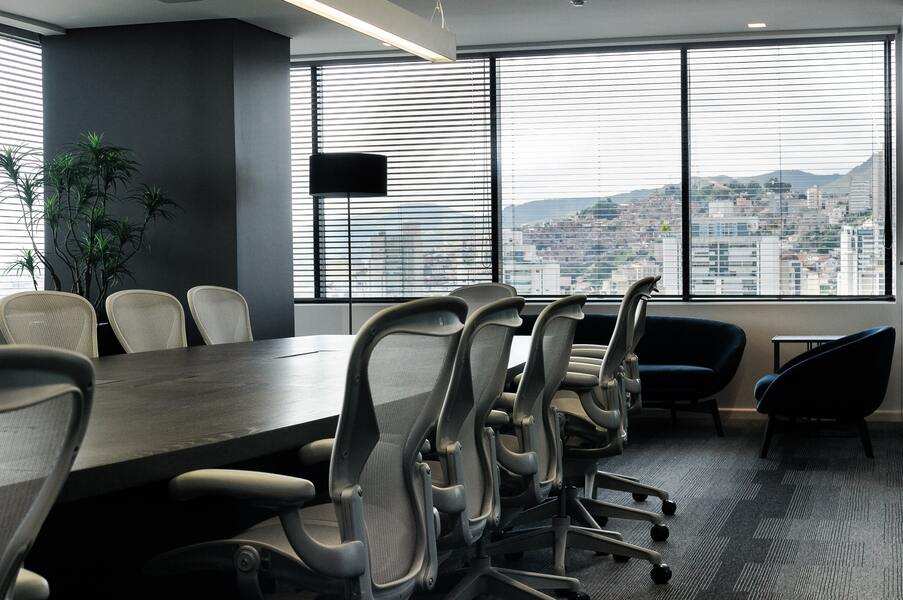 Una moderna sala de conferencias con una larga mesa, sillas de oficina ergonómicas y grandes ventanales con vistas a los edificios de la ciudad. Imagen alegórica sobre la movilidad geográfica centro de trabajo. Las macetas y los sillones de la esquina añaden un toque acogedor.