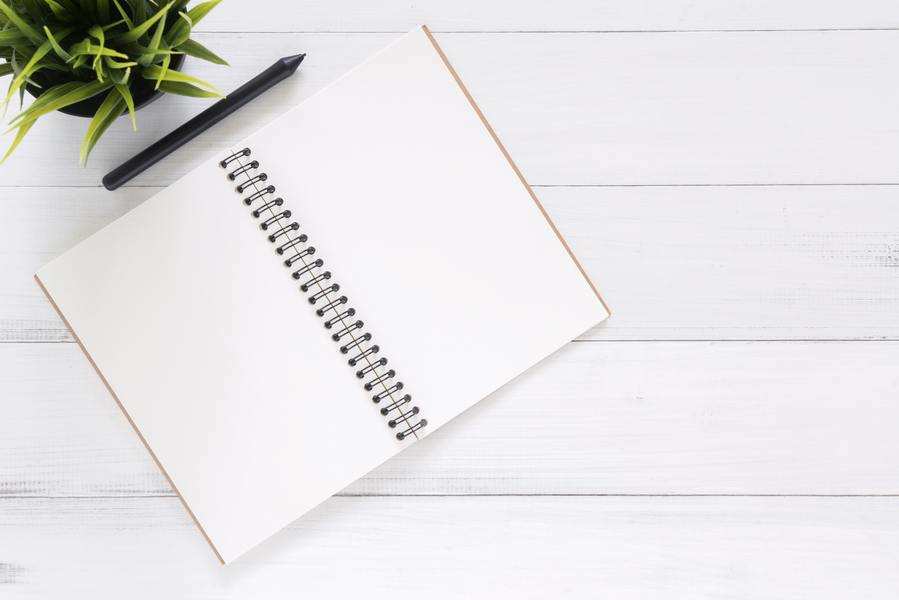 Un cuaderno de espiral abierto con páginas blancas en blanco descansa sobre una superficie de madera blanca, listo para tomar notas sobre movilidad geográfica centro de trabajo. Encima del cuaderno hay un bolígrafo negro y en la esquina superior izquierda se ve parcialmente una maceta verde.
