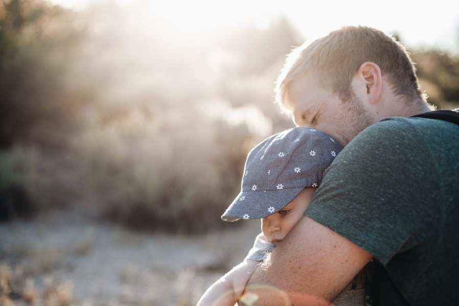 Un hombre abraza a un niño pequeño al aire libre durante el día, reflejando la cercanía que suele verse en una reclamación de paternidad. El niño lleva una gorra azul con dibujos blancos, mientras que la luz del sol crea un fondo cálido y suave. Ambos parecen tranquilos y conectados.