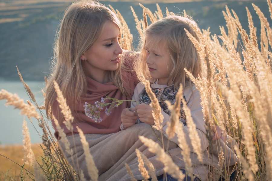 Una mujer y una niña se sientan juntas en un campo de hierba alta y seca, la mujer envuelta en una manta y sosteniendo flores. Se miran atentamente, compartiendo un momento tranquilo, como si contemplaran los cambios de la vida como testamento o divorcio. El fondo está difuminado con un paisaje natural.