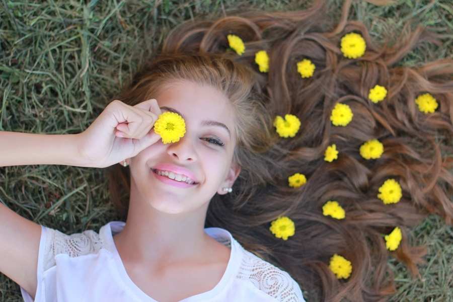 Una joven está tumbada sobre la hierba, sonriente, con su larga melena esparcida a su alrededor. Sostiene una flor amarilla sobre un ojo: su alegría despreocupada hace que preocupaciones como el carnet conducir gasto extraordinario parezcan lejanas. Lleva una camisa blanca y flores en el pelo.