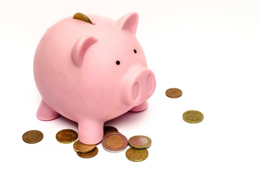 Una hucha rosa con una moneda introducida en su ranura está rodeada de monedas esparcidas, que simbolizan cómo el ahorro puede ayudar a gestionar cualquier gasto extraordinario, como los gastos del carné de conducir, sobre un fondo blanco.