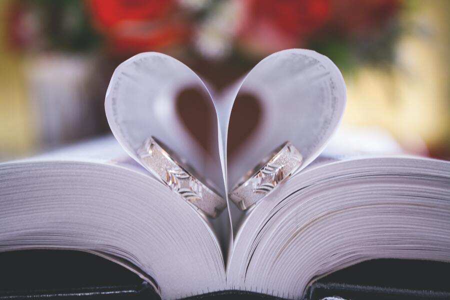 Un libro abierto con sus páginas dobladas hacia dentro para formar un corazón simboliza el amor y las capitulaciones matrimoniales. Dos anillos descansan en el interior del corazón, mientras que el fondo difuminado en rojo y verde añade un toque romántico. Un libro abierto con sus páginas dobladas hacia dentro para formar un corazón simboliza el amor y las capitulaciones matrimoniales. Dos anillos descansan en el interior del corazón, mientras que el fondo difuminado en rojo y verde añade un toque romántico.