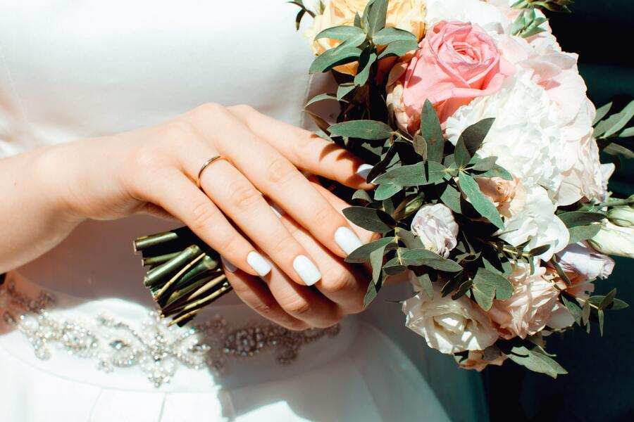 Primer plano de una persona vestida de blanco que sostiene un ramo de flores, luce un anillo de oro -quizá tras firmar unas capitulaciones matrimoniales- y unas uñas blancas pulcramente cuidadas. El ramo lleva rosas rosas y blancas con hojas verdes.