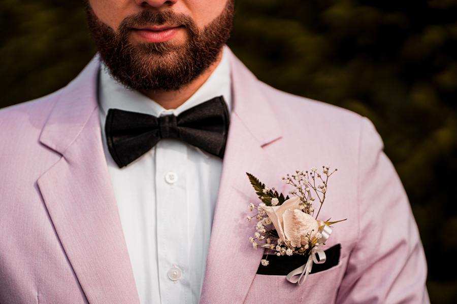 Un hombre vestido con traje de chaqueta rosa claro, camisa blanca y pajarita negra, con una flor en el ojal hecha con una rosa blanca y aliento de bebé en el bolsillo de la chaqueta, posa pensativo -quizá reflexionando sobre las próximas capitulaciones matrimoniales- frente a una vegetación borrosa.