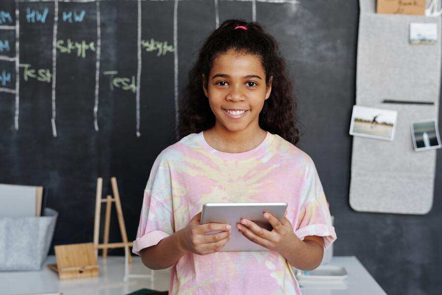 Una joven de pelo rizado, con una camiseta teñida, sujeta una tableta. Sonríe, y detrás de ella hay una pizarra con dibujos y gráficos, tal vez debatiendo temas como cambiar de escuela a un hijo sin autorización.