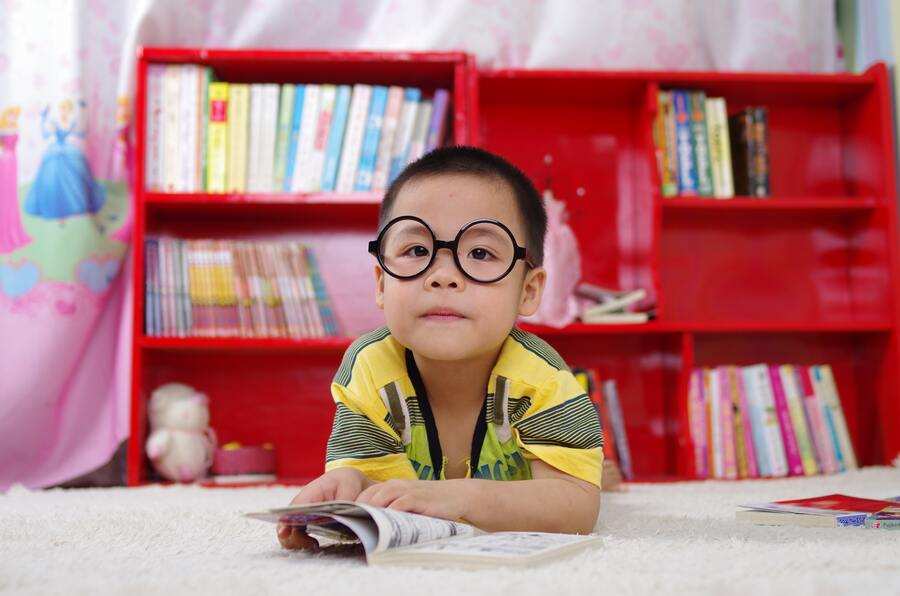 Un niño pequeño con gafas redondas y camisa amarilla está tumbado en una alfombra blanca con un libro abierto en la mano. Detrás del niño hay una estantería roja llena de libros y algunos juguetes: una escena acogedora que podría cambiar si cambias de escuela hijo sin autorización.