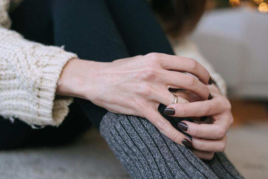 Una persona con jersey de punto color crema y pantalones oscuros se sienta con las rodillas levantadas, las manos juntas sobre un calcetín gris, mostrando el esmalte de uñas oscuro y un simple anillo, captando un momento tranquilo que puede insinuar los efectos del maltrato psicológico.