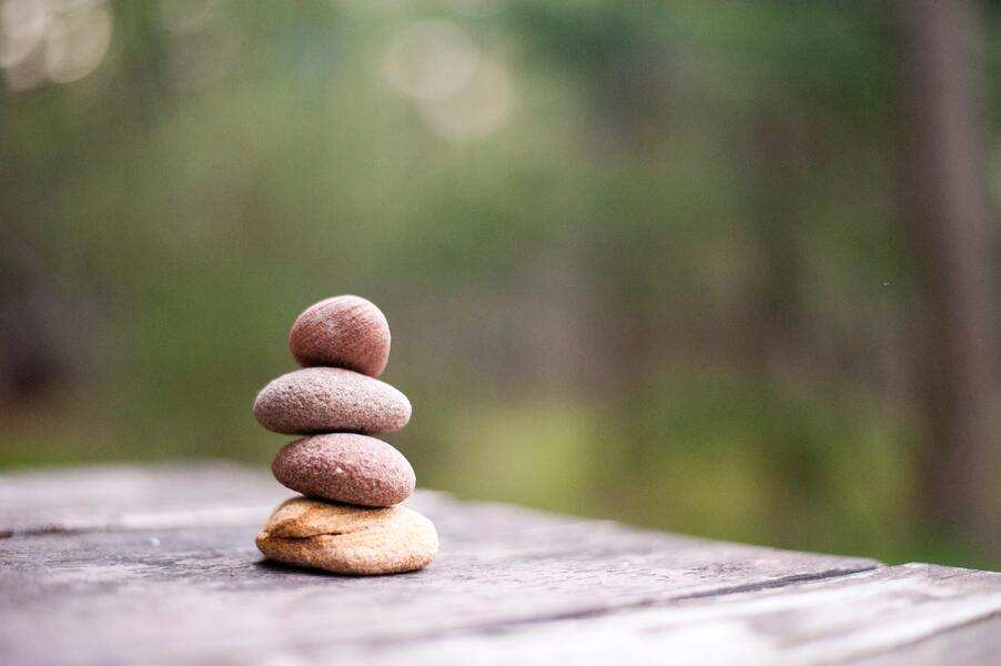 Sobre una superficie de madera se apilan cuatro piedras lisas que simbolizan el equilibrio y la resistencia, sobre un fondo natural difuminado en verde y marrón, una imagen que evoca la fortaleza frente a retos como el maltrato psicológico.