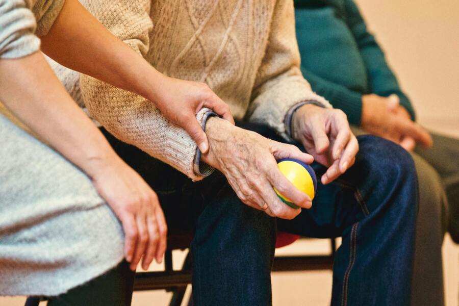 Una persona con jersey sostiene una pequeña pelota amarilla y azul mientras otra le apoya suavemente el brazo, posiblemente en un internamiento no voluntario; una tercera persona está sentada cerca, parcialmente visible.