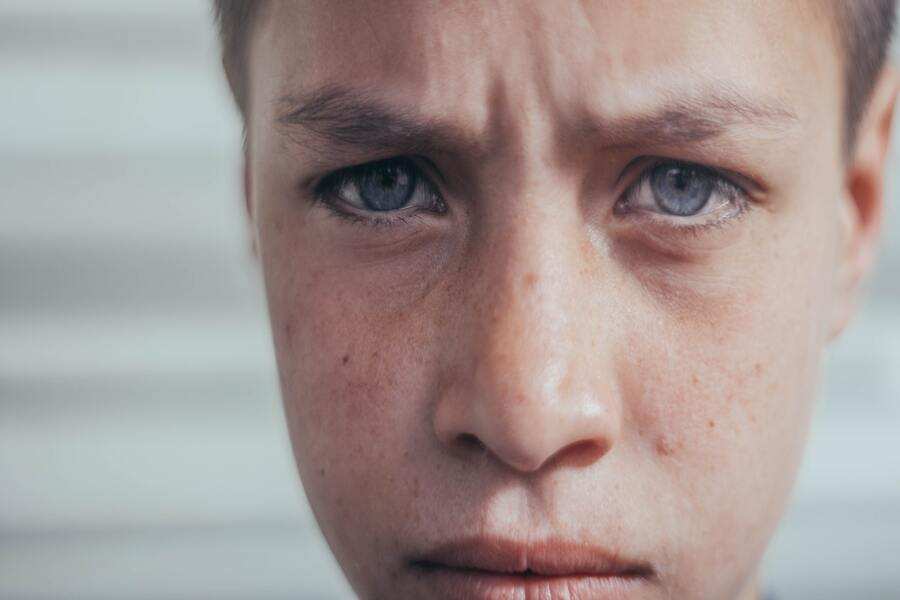 Primer plano del rostro de un joven con ojos azules y expresión seria o preocupada, frunciendo el ceño, que refleja la carga emocional del acoso escolar. El fondo es borroso y de color neutro.