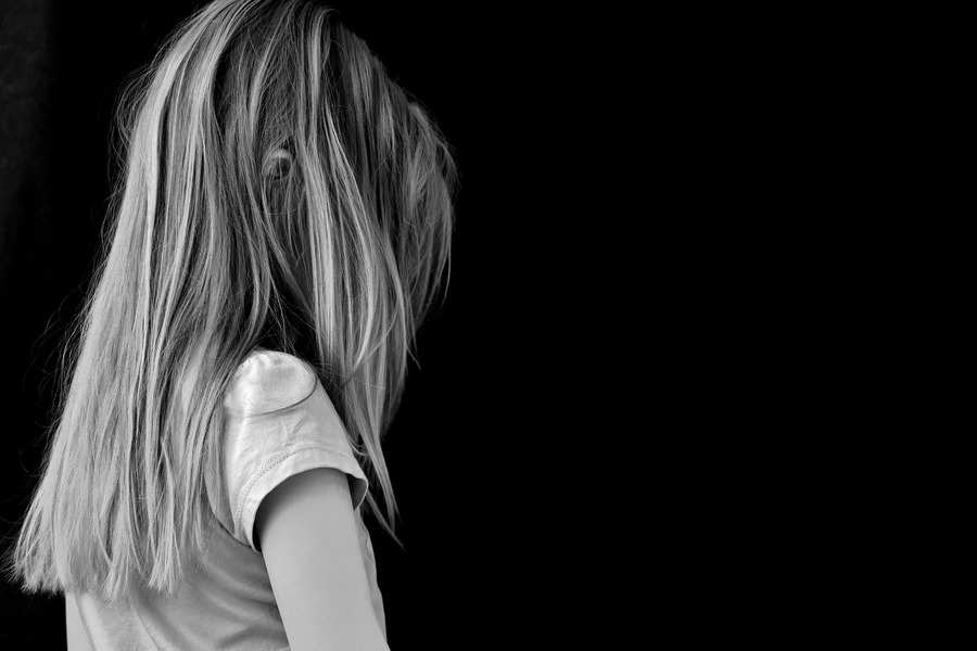 Una joven con el pelo largo y liso mira de espaldas a la cámara sobre un fondo negro liso, evocando el aislamiento que a menudo sienten las víctimas del acoso escolar. Lleva una camiseta de manga corta. No se le ve la cara.