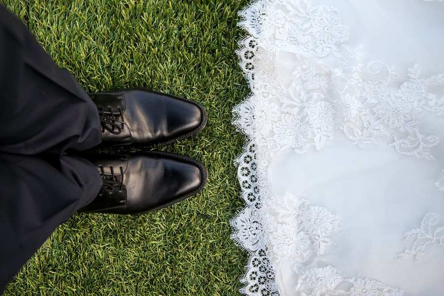 Primer plano de una persona con zapatos de vestir negros y pantalones negros de pie sobre la hierba junto al borde de un vestido de novia de encaje blanco, símbolo de la unión en sociedad de gananciales.