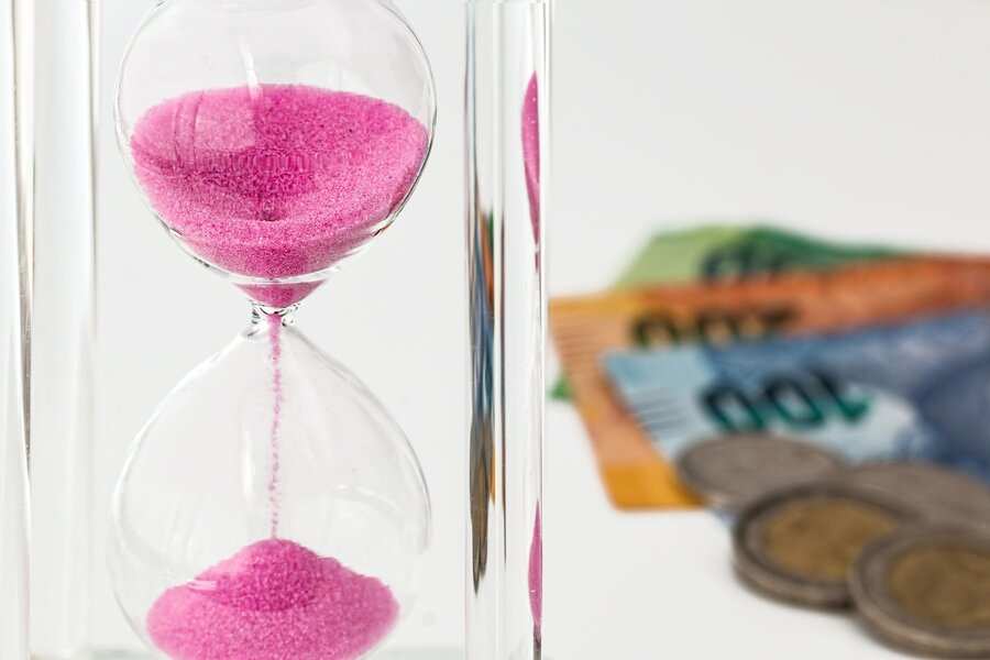 En primer plano aparece un reloj de arena transparente con arena rosa, que simboliza el paso del tiempo en una sociedad de gananciales, mientras que en el fondo se ven borrosos varios billetes y monedas sobre una superficie blanca.