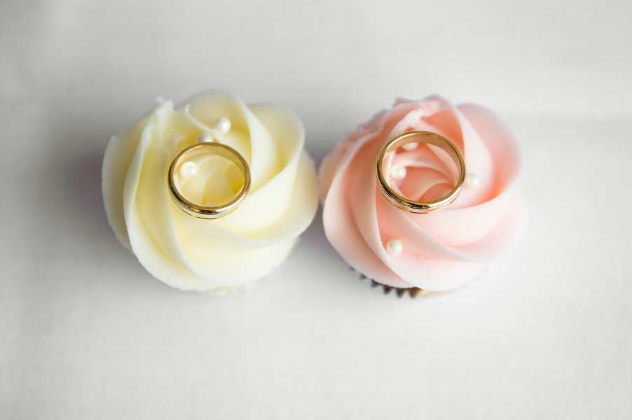 Dos magdalenas, una con glaseado blanco y otra con glaseado rosa, cada una coronada con un anillo dorado y una pequeña perla blanca decorativa, se colocan una al lado de la otra sobre un fondo blanco, simbolizando la unidad como sociedad de gananciales. Dos magdalenas, una con glaseado blanco y otra con glaseado rosa, cada una coronada con un anillo dorado y una pequeña perla blanca decorativa, se colocan una al lado de la otra sobre un fondo blanco, simbolizando la unidad como sociedad de gananciales.