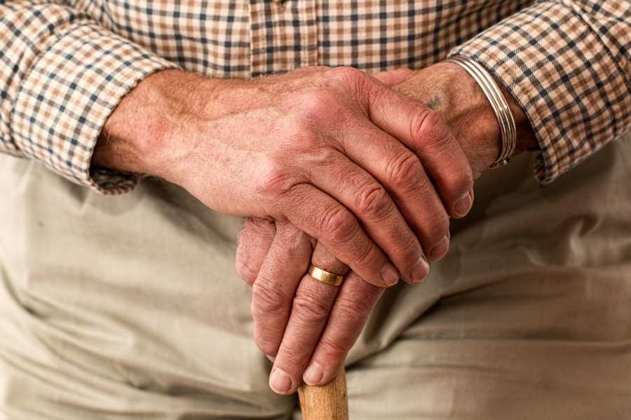 Primer plano de las manos de un adulto mayor apoyadas en un bastón de madera, con un anillo de oro y un brazalete de metal -símbolos de vida y legado, a menudo tenidos en cuenta a la hora de crear un testamento vital-, con una camisa a cuadros y unos pantalones beige de fondo.