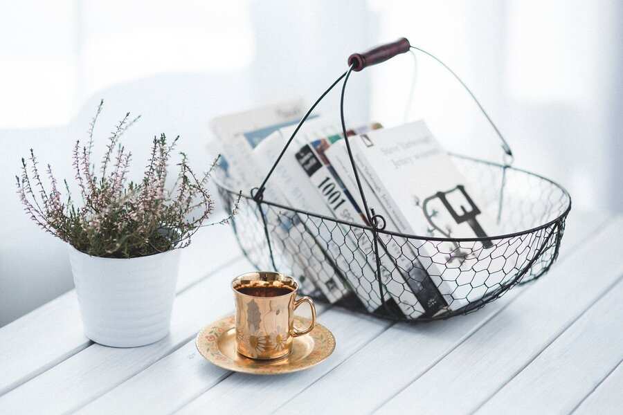 Una cesta de alambre sostiene varios libros y periódicos sobre una mesa de madera blanca junto a una maceta y una taza de té dorada con su platillo a juego, tal vez inspirando pensamientos sobre codicilo memorias testamentarias o cómo modificar testamento. El fondo está suavemente iluminado. Una cesta de alambre sostiene varios libros y periódicos sobre una mesa de madera blanca junto a una maceta y una taza de té dorada con su platillo a juego, tal vez inspirando pensamientos sobre codicilo memorias testamentarias o cómo modificar testamento. El fondo está suavemente iluminado.